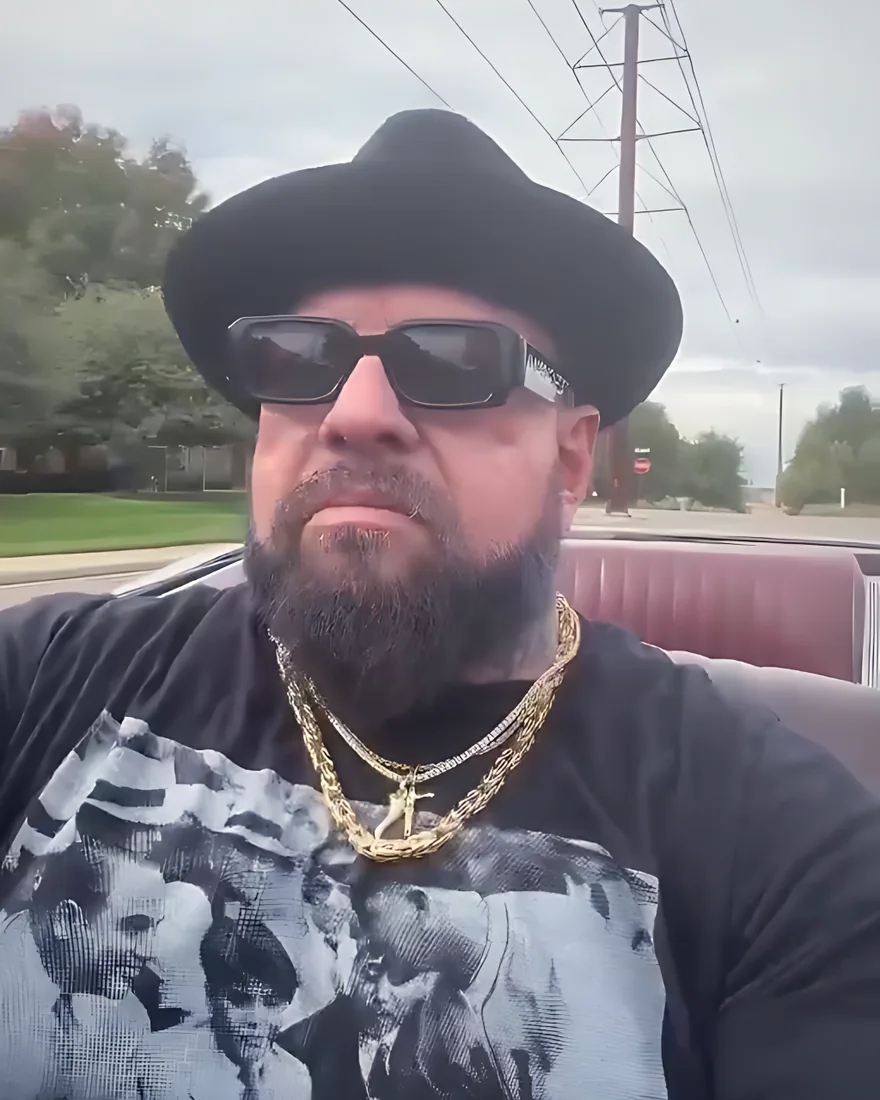 Face behind the wheel of his convertible, in a black hat, sunglasses and gold chains.