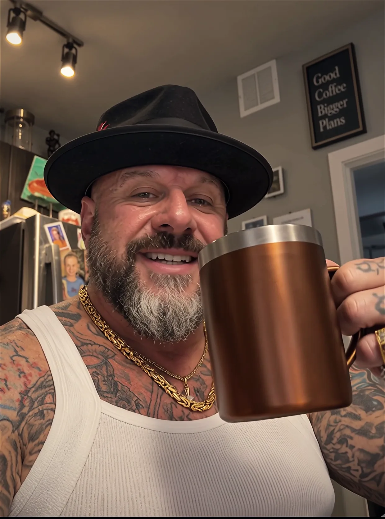 Face lowering the copper mug and smiling into the camera, with the Good Coffee, Bigger Plans sign on his kitchen wall behind him.